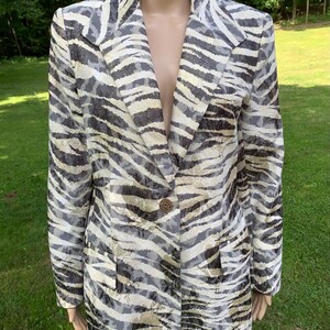 May include: A long-sleeved blazer with a zebra print pattern in shades of black, white, and gold. The blazer has a single button closure and two pockets.