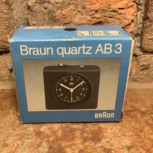May include: A black Braun quartz AB 3 alarm clock in its original box. The clock has a black face with white numbers and hands. The box is blue with white text.