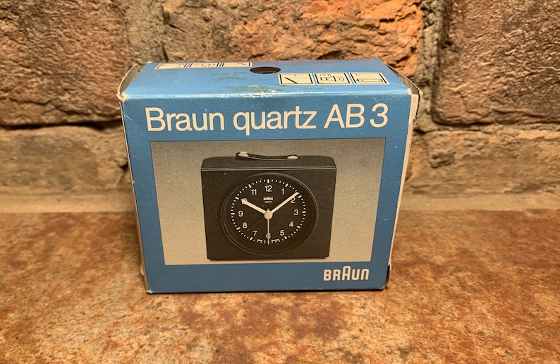 May include: A black Braun quartz AB 3 alarm clock in its original box. The clock has a black face with white numbers and hands. The box is blue with white text.