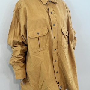 May include: Mustard-colored button-down shirt with two chest pockets. The shirt has a collared neckline and long sleeves. The fabric appears to be a durable, workwear-style material. The shirt is displayed on a mannequin.