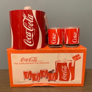 May include: A vintage Coca-Cola brand ice bucket and on-the-rocks set. The set includes a red and white ice bucket, and three glasses with the Coca-Cola logo. The set is in its original packaging.