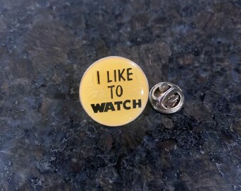 May include: A yellow enamel pin with the text "I like to watch" in black lettering.
