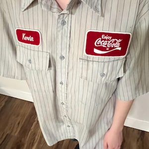 May include: A light gray, short-sleeved work shirt with vertical black pinstripes. The shirt features two red rectangular patches: one with the name "Kevin" and the other with the Coca-Cola logo and the words "Enjoy Coca-Cola".