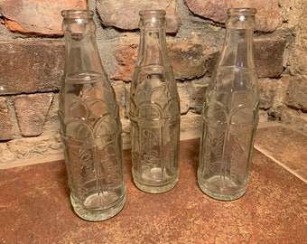 May include: Three clear glass bottles with embossed designs. The bottles are empty and have a vintage look.