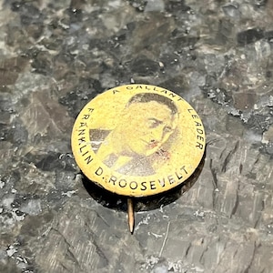 May include: A vintage, round pin featuring a portrait of Franklin D. Roosevelt. The pin has a yellowed background with the text "A GALLANT LEADER" and "FRANKLIN D. ROOSEVELT" encircling the image. A straight pin extends from the back.