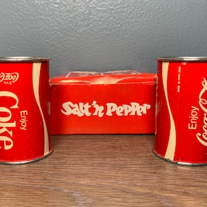 May include: A set of vintage Coca-Cola themed items. Two cylindrical Coke cans flank a red box labeled "Salt 'n Pepper". The cans are red with white Coca-Cola branding and the words "Enjoy Coke".