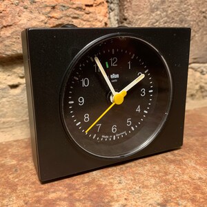 May include: A black alarm clock with a white face and yellow hands. The clock reads "Elan Uhr" and "Made in Germany".