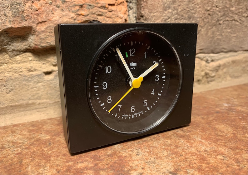 May include: A black alarm clock with a white face and yellow hands. The clock reads "Elan Uhr" and "Made in Germany".