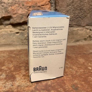 May include: A white box with blue accents and the Braun logo. The box contains a battery-operated alarm clock. The text on the box reads "Batteriewecker (1,5V Mignonzelle). Leicht zu bedienen. Angenehmes Wecksignal in Intervallen. Unzerbrechliches Geh&auml;use. 1 Jahr Garantie." and "Battery alarm clock (1,5 V mignon cell). Easy operation. Clear intermittent alarm signal, shatterproof housing. 1 year guarantee."