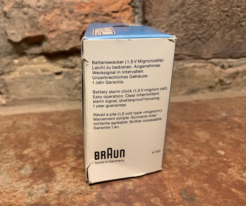 May include: A white box with blue accents and the Braun logo. The box contains a battery-operated alarm clock. The text on the box reads "Batteriewecker (1,5V Mignonzelle). Leicht zu bedienen. Angenehmes Wecksignal in Intervallen. Unzerbrechliches Geh&auml;use. 1 Jahr Garantie." and "Battery alarm clock (1,5 V mignon cell). Easy operation. Clear intermittent alarm signal, shatterproof housing. 1 year guarantee."
