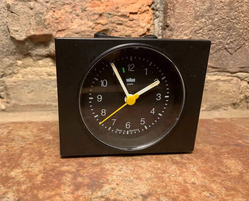 May include: A black alarm clock with a white face and black hands. The clock has a yellow second hand and the words "Braun quartz" printed on the face. The clock is made in Germany.