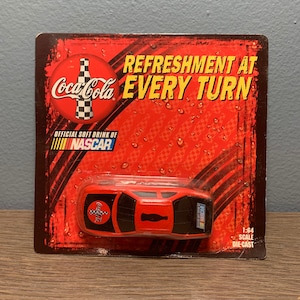 Op de afbeelding: Een gegoten modelauto in een blisterverpakking. De auto is rood en zwart met Coca-Cola-branding en NASCAR-logo's. De verpakking is rood met het Coca-Cola-logo en de tekst "REFRESHMENT AT EVERY TURN". Schaal 1:64.