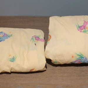 May include: Two yellow fitted sheets with a cartoon animal print. The sheets feature a light yellow background with a variety of colorful cartoon animals, including a blue bunny, a pink bunny, and a yellow bird.