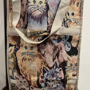 Könnte beinhalten: Eine beige Tragetasche mit einem Tapisserie-Design, das verschiedene Katzen in verschiedenen Farben und Posen zeigt. Die Tasche hat zwei Henkel und einen Reißverschluss.