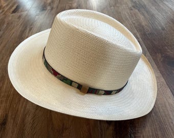 May include: A wide-brimmed straw hat with a woven band in shades of green, red, and black. The hat is made of natural straw and has a light beige color.