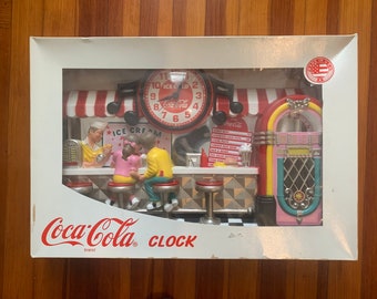 May include: A Coca-Cola branded clock featuring a vintage diner scene with a jukebox, a counter, and a clock face with the Coca-Cola logo. The clock is framed in a white box with the words "Coca-Cola brand CLOCK" printed on the bottom.