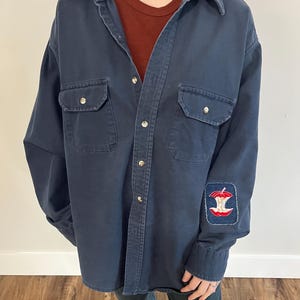 May include: A dark blue long-sleeved work shirt with a button-up front and two chest pockets. The shirt has a red, white and blue apple patch on the left sleeve.