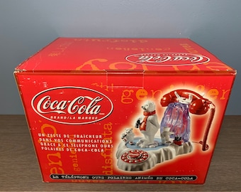 May include: A red Coca-Cola branded box with a white polar bear and a red rotary phone on a snowy landscape. The box reads "Coca-Cola Brand/La Marque" and "Un Zeste de Fraicheur Dans Vos Communications Grace a Ce Telephone Ours Polaires de Coca-Cola". The box also reads "Le Telephone Ours Polaires Animes de Coca-Cola".