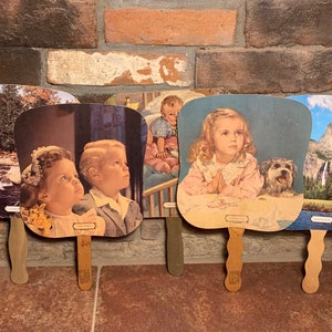 Puede incluir: Cuatro abanicos de papel vintage con diferentes imágenes. Un abanico presenta una escena de cascada, otro tiene una imagen de dos niños, un tercero tiene una niña rezando con un perro y el último muestra un paisaje de montaña.