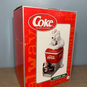 May include: A red and white Coca-Cola cookie jar featuring a polar bear wearing a Coca-Cola hat and holding a bottle of Coca-Cola. A penguin is standing next to the polar bear. The box is red and white and says "Coke" and "always".