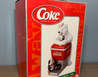 May include: A red and white Coca-Cola cookie jar featuring a polar bear wearing a Coca-Cola hat and holding a bottle of Coca-Cola. A penguin is standing next to the polar bear. The box is red and white and says "Coke" and "always".