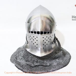 Puede incluir: Un casco plateado con una parte superior puntiaguda y una máscara facial con agujeros. El casco se lleva sobre una cota de malla. El texto "Honour Kraft Medieval+Hand+Forged+Armor" es visible en la esquina superior derecha de la imagen.
