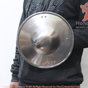 May include: A silver metal shield with a domed center and a leather strap. The shield is held by a person wearing a black sweatshirt.