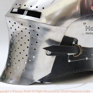 May include: A silver helmet with a face mask and a black leather strap. The helmet has a series of small holes throughout the face mask. The helmet is made by Honour Kraft, a company that specializes in medieval hand-forged armor.