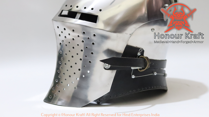 May include: A silver helmet with a face mask and a black leather strap. The helmet has a series of small holes throughout the face mask. The helmet is made by Honour Kraft, a company that specializes in medieval hand-forged armor.