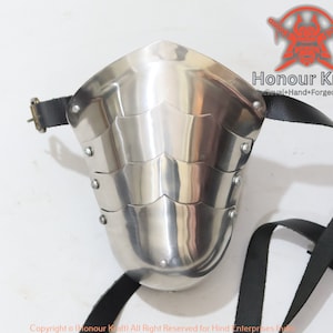 Könnte beinhalten: Ein silberner Schienbeinschoner mit Schuppenmuster und einem schwarzen Lederriemen. Der Schienbeinschoner ist aus Metall und hat eine gebogene Form. Der Riemen ist mit Metallnieten am Schienbeinschoner befestigt. Der Schienbeinschoner ist ein Teil einer mittelalterlichen Rüstung.