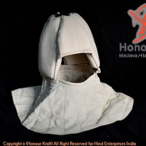 Puede incluir: Un casco acolchado blanco con capucha, diseñado para protección durante el combate medieval. El casco está hecho de tela gruesa y tiene una forma redondeada. Está diseñado para cubrir la cabeza y el cuello.
