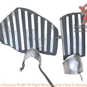 May include: A pair of medieval-style leg armor made of metal plates and leather straps. The armor is silver and black and has a striped pattern. The armor is attached to the legs with leather straps. The armor is for sale on the website Honour Kraft.