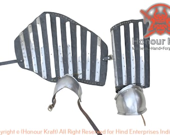 May include: A pair of medieval-style leg armor made of metal plates and leather straps. The armor is silver and black and has a striped pattern. The armor is attached to the legs with leather straps. The armor is for sale on the website Honour Kraft.