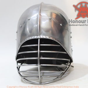 Op de afbeelding: Een zilveren helm met een gezichtsmasker gemaakt van metalen staven. De helm heeft een afgeronde bovenkant en een puntige kinbeschermer. De helm is gemaakt van metaal en heeft een zilveren afwerking.