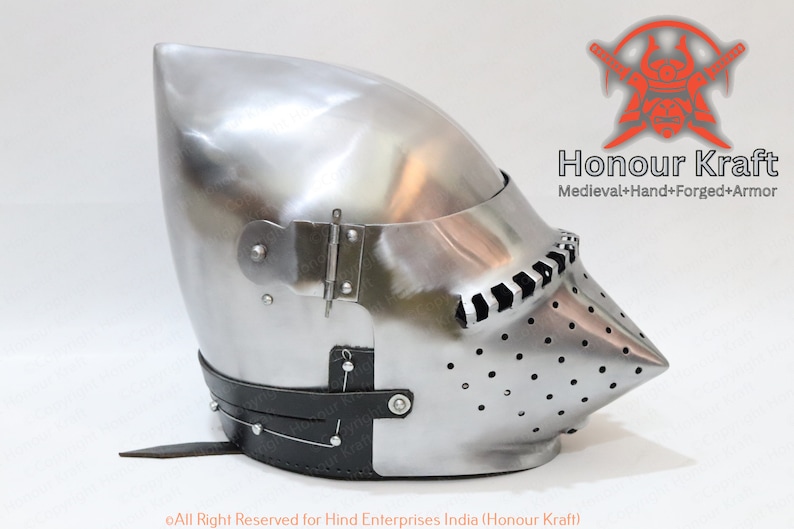 K&ouml;nnte beinhalten: Ein silberner Helm mit einem spitzen Visier und einem Lederriemen. Der Helm hat ein Scharnier-Visier und eine Reihe kleiner L&ouml;cher &uuml;ber die gesamte Oberfl&auml;che. Der Helm wird von Honour Kraft hergestellt, einem Unternehmen, das sich auf handgeschmiedete mittelalterliche R&uuml;stungen spezialisiert hat.