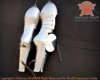 May include: A pair of silver metal knee armor, hanging from hooks on a black background. The armor is made of metal plates and has a smooth, polished finish. The armor is designed to protect the knees and shins.