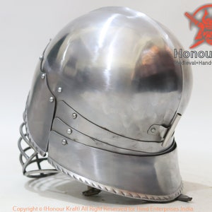 Op de afbeelding: Een zilveren helm met een gezichtsbeschermer en een decoratieve rand. De helm is gemaakt van metaal en heeft een gladde, gepolijste afwerking.