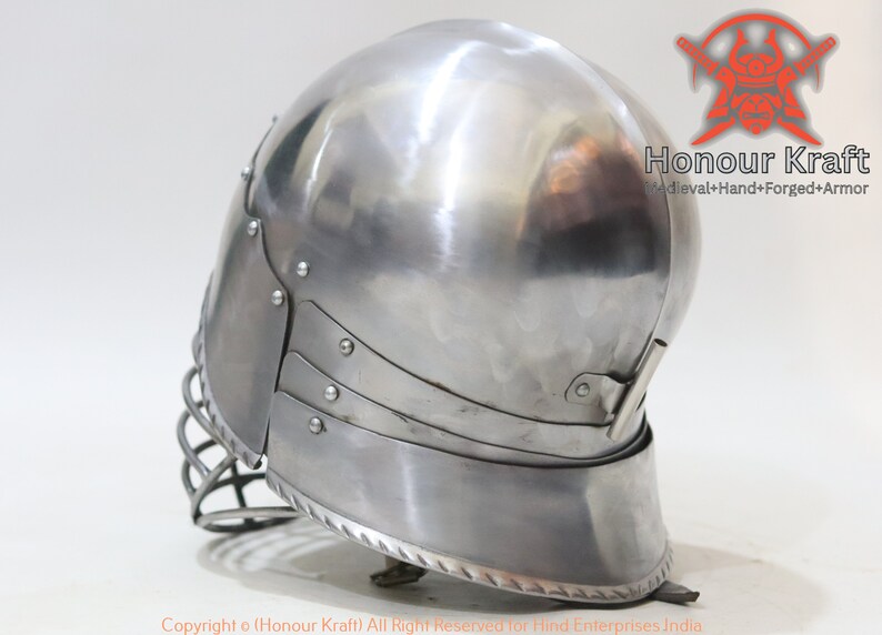 Op de afbeelding: Een zilveren helm met een gezichtsbeschermer en een decoratieve rand. De helm is gemaakt van metaal en heeft een gladde, gepolijste afwerking.