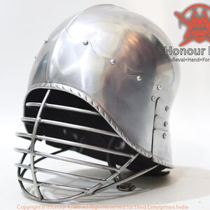 Op de afbeelding: Een zilveren helm met een gezichtskooi. De helm heeft een afgeronde bovenkant en een vizier dat de ogen bedekt. De kooi is gemaakt van metalen staven en is met klinknagels aan de helm bevestigd.