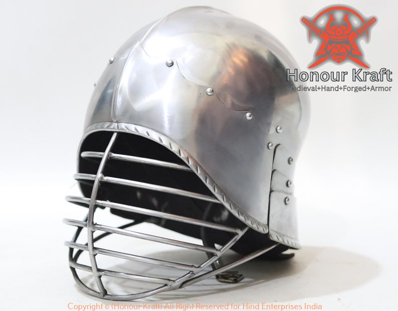 Op de afbeelding: Een zilveren helm met een gezichtskooi. De helm heeft een afgeronde bovenkant en een vizier dat de ogen bedekt. De kooi is gemaakt van metalen staven en is met klinknagels aan de helm bevestigd.