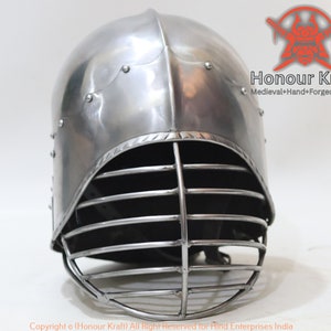 Op de afbeelding: Een zilveren helm met een metalen gezichtsmasker. De helm heeft een afgeronde bovenkant en een vizier dat de ogen bedekt. Het gezichtsmasker is gemaakt van metalen staven die uit elkaar staan om zicht te garanderen. De helm is een helm in middeleeuwse stijl.