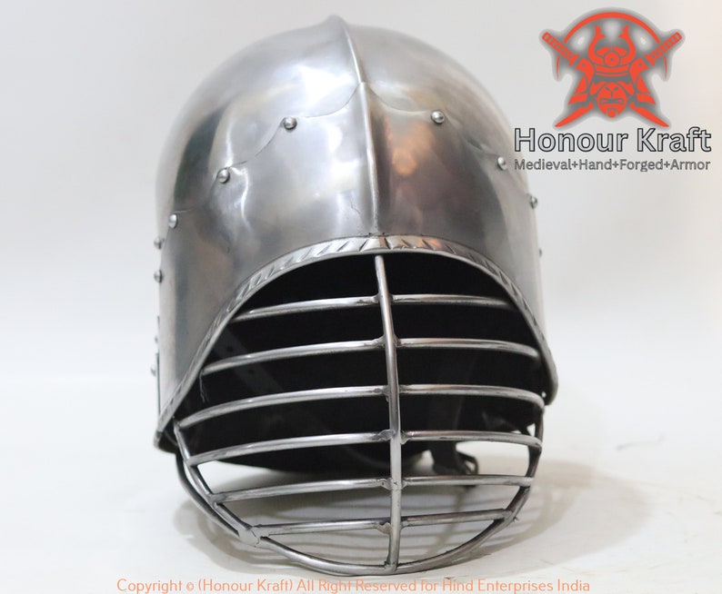Op de afbeelding: Een zilveren helm met een metalen gezichtsmasker. De helm heeft een afgeronde bovenkant en een vizier dat de ogen bedekt. Het gezichtsmasker is gemaakt van metalen staven die uit elkaar staan om zicht te garanderen. De helm is een helm in middeleeuwse stijl.