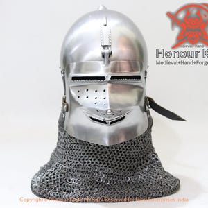 May include: A polished silver medieval helmet with a visor and a chainmail neck guard. The helmet has a hinged visor with eye slits and ventilation holes. The text "Honour Kraft" is visible on the right side of the image.