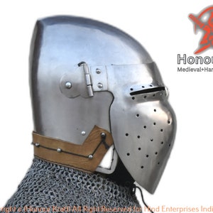 May include: A side view of a medieval helmet with a visor and a leather strap. The helmet is made of metal and has a textured surface. The helmet is a hand-forged piece of armour.