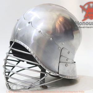 Op de afbeelding: Een zilveren helm met een gezichtsbeschermer, gemaakt van metaal en handgesmeed. De helm heeft een afgeronde bovenkant en een vizier dat het gezicht bedekt. De helm is een helm in middeleeuwse stijl en is een onderdeel van een harnas.