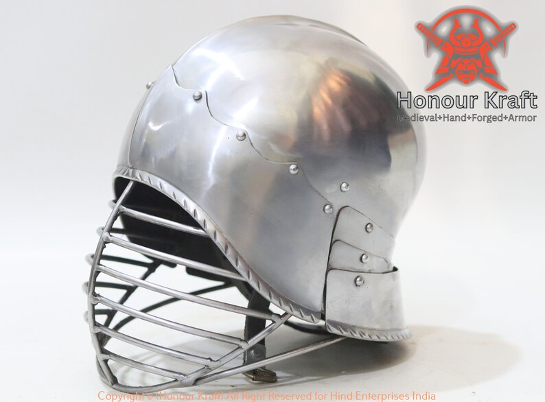 Op de afbeelding: Een zilveren helm met een gezichtsbeschermer, gemaakt van metaal en handgesmeed. De helm heeft een afgeronde bovenkant en een vizier dat het gezicht bedekt. De helm is een helm in middeleeuwse stijl en is een onderdeel van een harnas.
