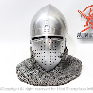 Puede incluir: Un casco medieval de plata con visera y cota de malla. El casco tiene una parte superior puntiaguda y una pequeña abertura rectangular para los ojos. La visera es de metal y tiene una serie de pequeños agujeros para la ventilación. La cota de malla está hecha de anillos de metal entrelazados y cubre el cuello y los hombros.