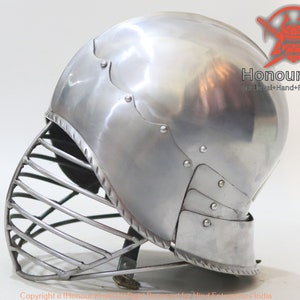 Op de afbeelding: Een zilveren helm met een gezichtsmasker van metalen staven. De helm heeft een afgeronde bovenkant en een puntige kinbeschermer. De helm is gemaakt van metaal en heeft een glanzende afwerking. De helm is een type middeleeuwse bepantsering.