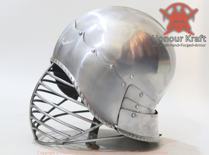 Op de afbeelding: Een zilveren helm met een gezichtsmasker van metalen staven. De helm heeft een afgeronde bovenkant en een puntige kinbeschermer. De helm is gemaakt van metaal en heeft een glanzende afwerking. De helm is een type middeleeuwse bepantsering.