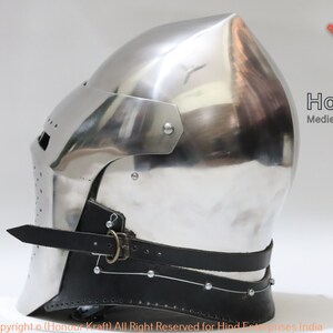 May include: A silver medieval helmet with a pointed top and a leather chin strap. The helmet is made of metal and has a textured surface. The helmet is a replica of a historical helmet.
